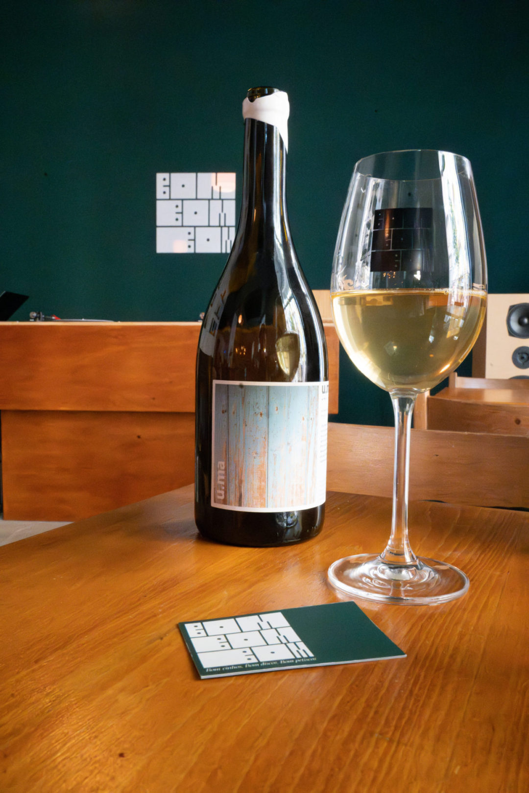 A glass of white wine next to an open bottle on a table in front of a DJ stand and a sign reading "BOM BOM BOM" at a natural wine bar in Lisbon, Portugal
