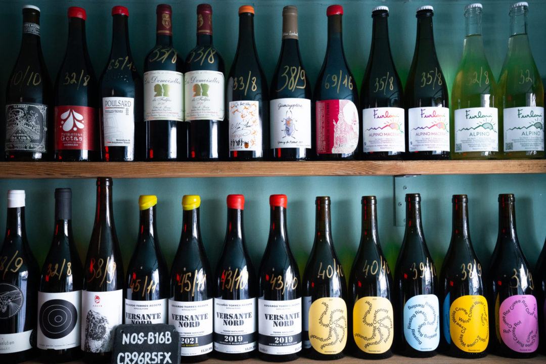 Two shelves full of bottled wines with prices by the bottle and by the glass listed on them inside Holy Wine!, one of the best spots for Natural Wine in Lisbon