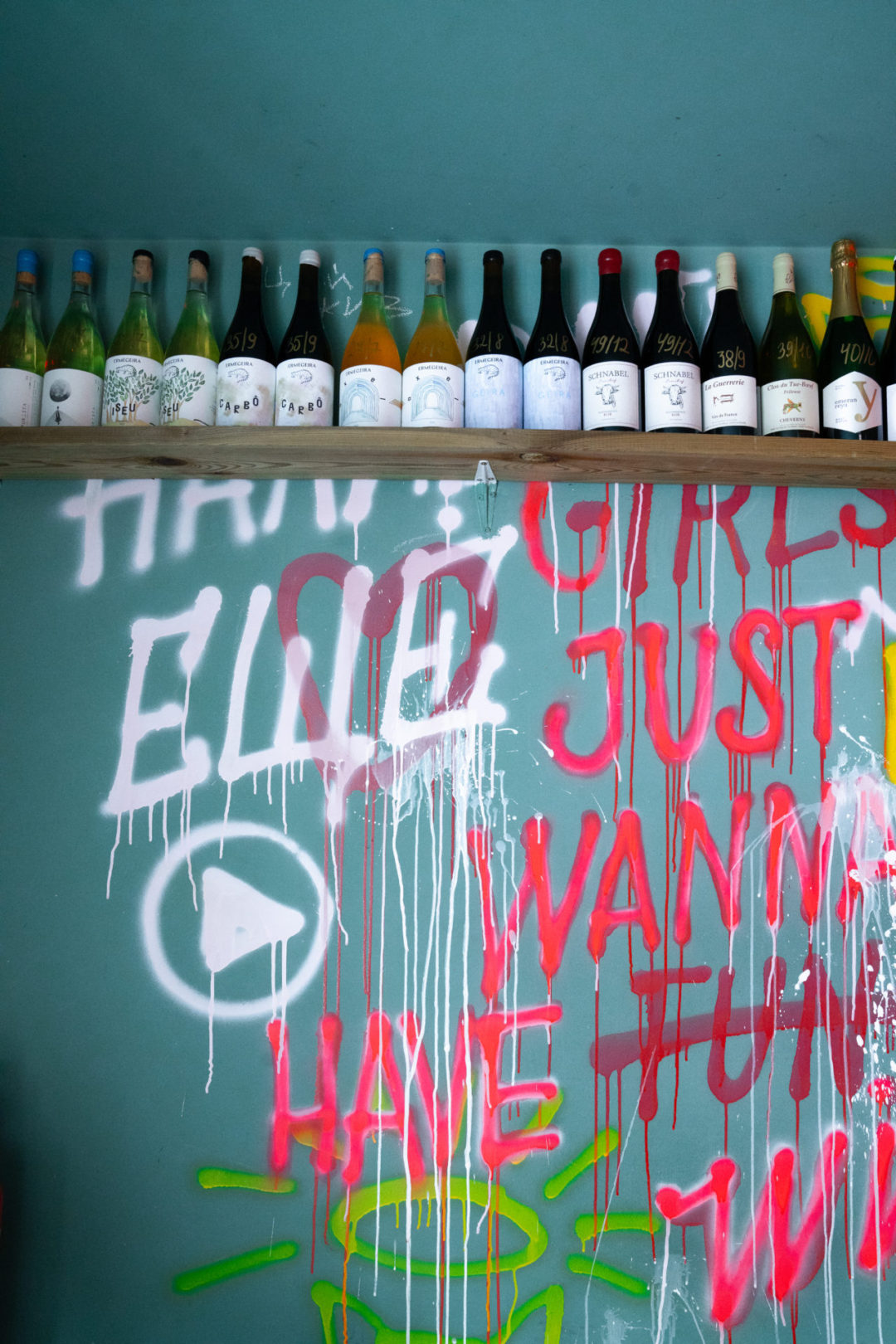 A shelf full of natural wines above a turqoise painted wall with graffiti "Girls Just Want to Have F̶u̶n̶ Wine" inside Holy Wine! in Lisbon