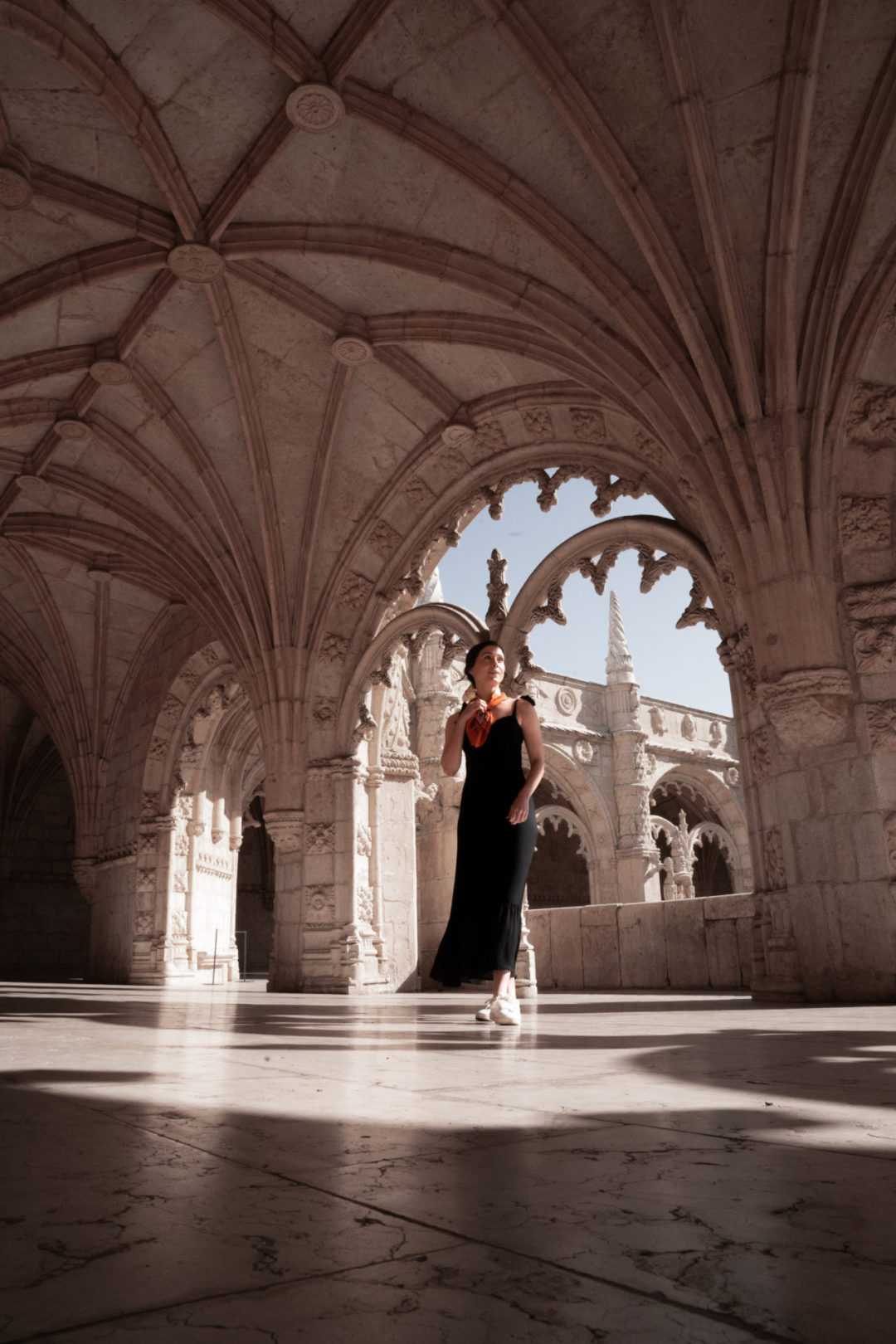 Travel Blogger Jordan Gassner wearing a black dress and orange silk scarf walking walking through Jerónimos Monastery in Lisbon