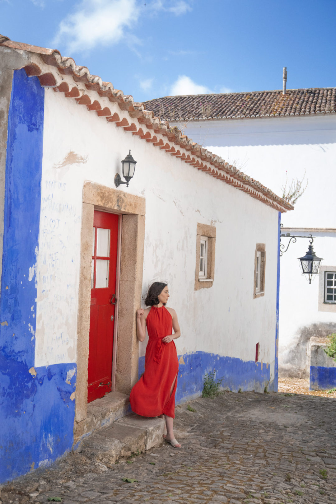 Travel Blogger Jordan Gassner leaning against a red doorway of a blue and white building while wearing a matching red dress in Obidos, Portugal
