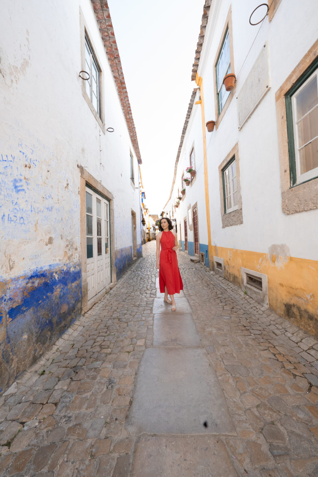Travel Blogger Jordan Gassner standing and smiling in the middle of Rua Direita next to bright yellow, blue and white shops in Obidos, Portugal