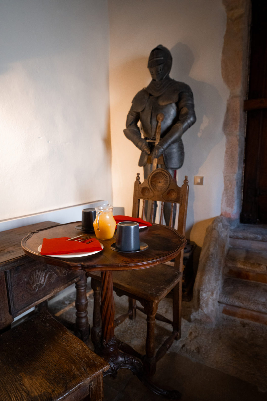 A glass of orange juice sitting on a table next to a suit of armor inside Torre De Maneys Hotel in Obidos, Portugal