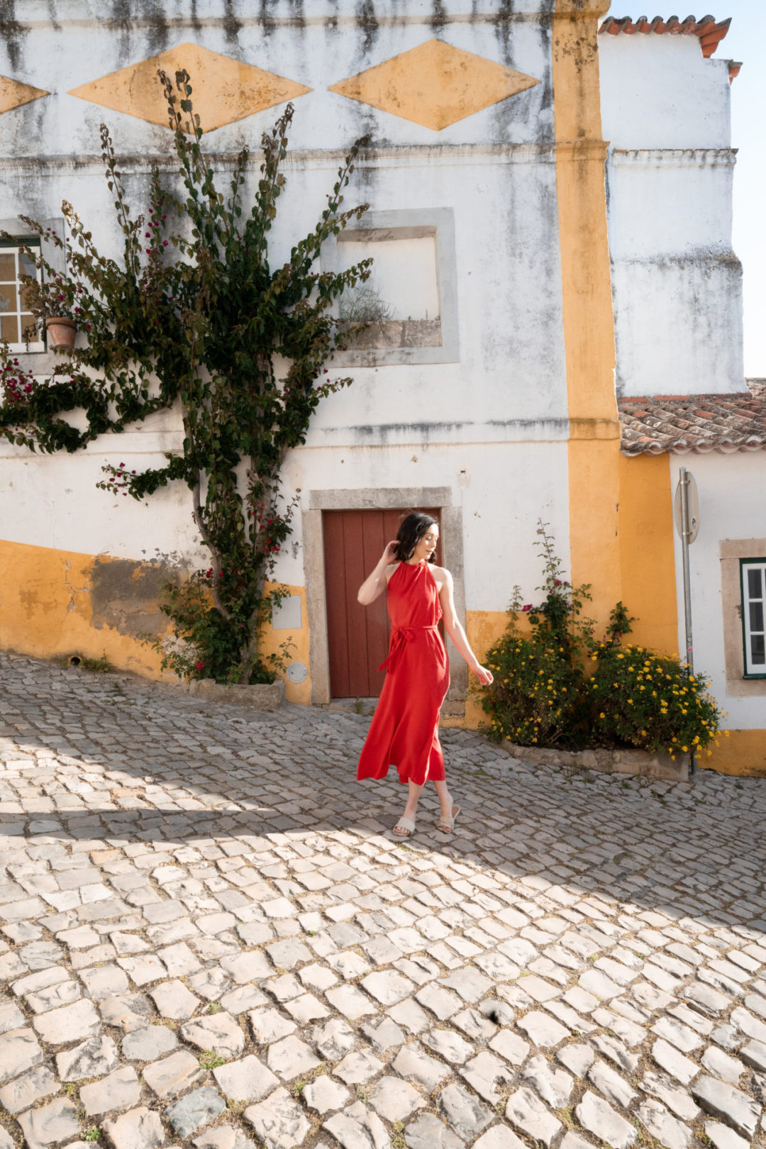 Travel Blogger Jordan Gassner wearing a bright red dress while dancing on a hill in front of a bright yellow and white home in Obidos, Portugal