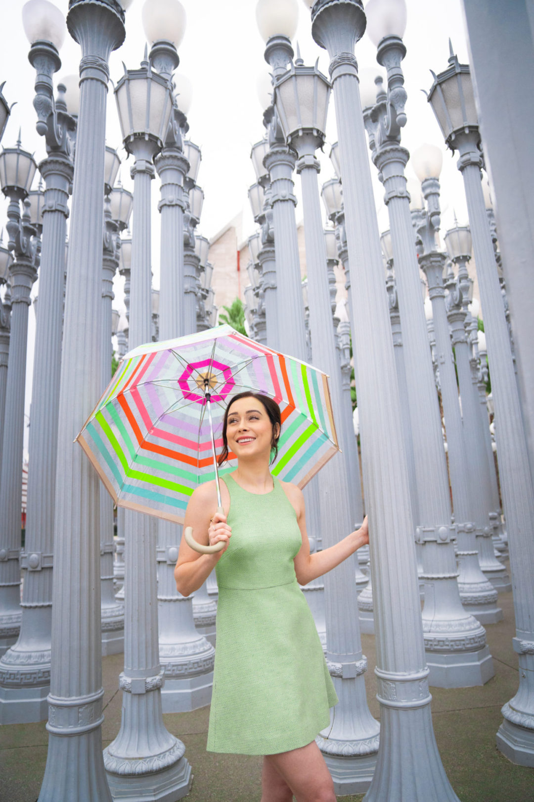 Travel Blogger Jordan Gassner smiling in a green dress under a rainbow umbrella near the "Urban Light" installation outside of LACMA