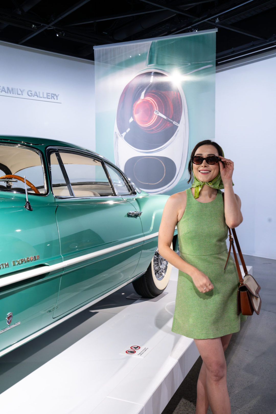 Travel Blogger Jordan Gassner wearing a a mint green dress and driving scarf next to a matching car, a 1954 Plymouth Explorer, at Petersen Automotive Museum in Los Angeles, California