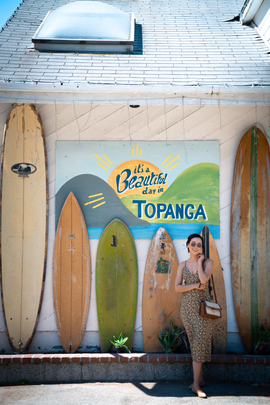 Travel Blogger Jordan Gassner smiling next to a "It's A Beautiful Day in Topanga" mural next to a set of 6 standing surboards inside a Topanga Canyon shopping center