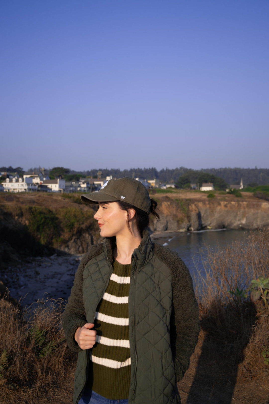 Travel Blogger Jordan Gassner looking over her shoulder and smiling along the cliffside in front of the town of Mendocino, California
