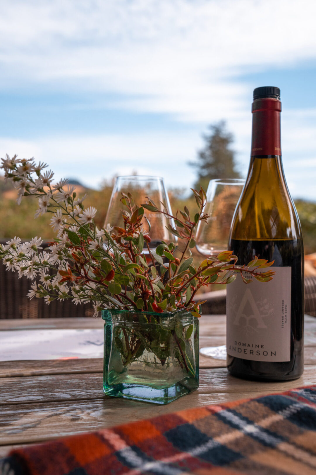 A bottle of wine next to two wine glasses and a vase of flowers on a table near a vineyard in Philo, California