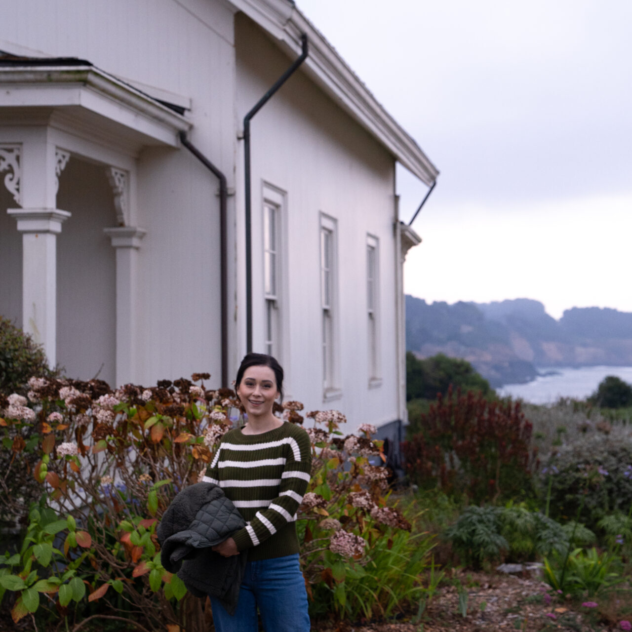 Travel Blogger Jordan Gassner smiling and holding her coat as sunrise approaches at the historic Ford House in Mendocino, California
