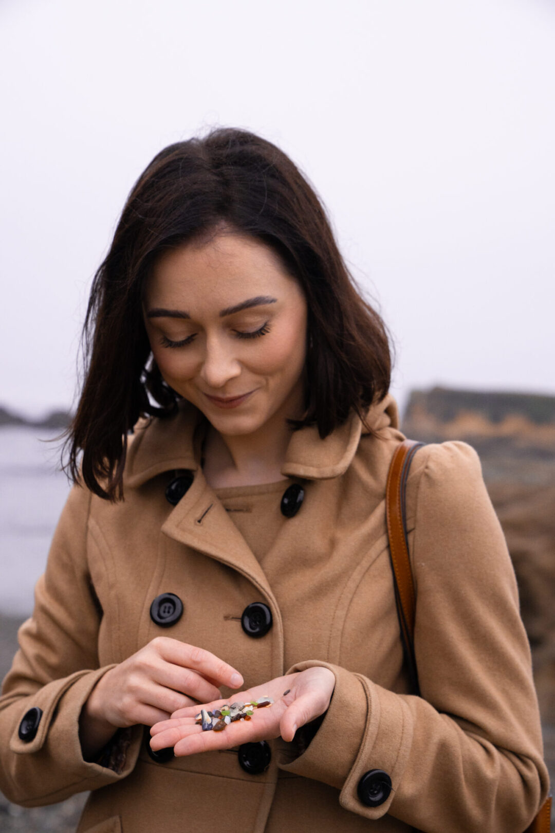 Travel Blogger Jordan Gassner smiling while looking down at some glass-sand in her hand from Glass Beach in Fort Bragg, California