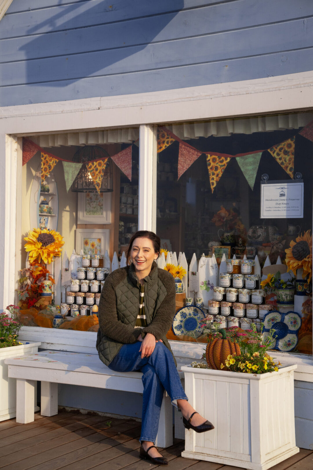Travel Blogger Jordan Gassner sitting on a white painted bench in front of the window of a blue Victorian building filled with jams and preserves