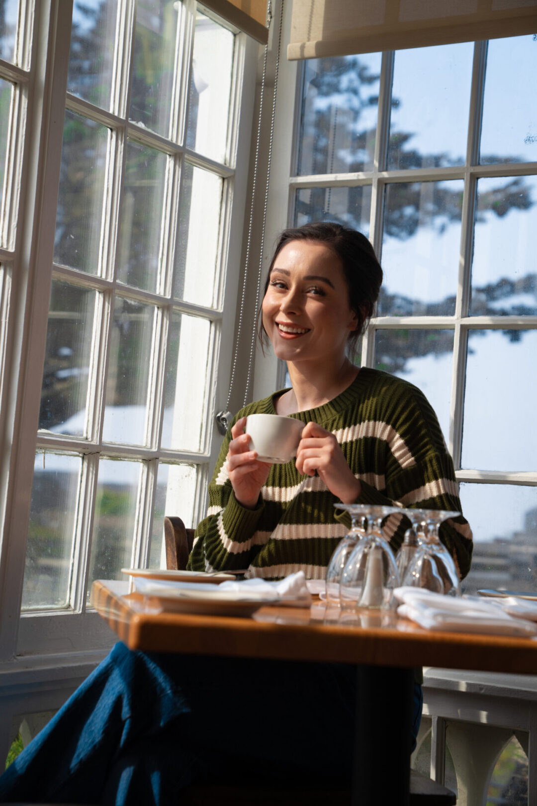 Travel Blogger Jordan Gassner smiling while holding a cup of tea in front of a Victorian era window inside MacCallum House in Mendocino, California
