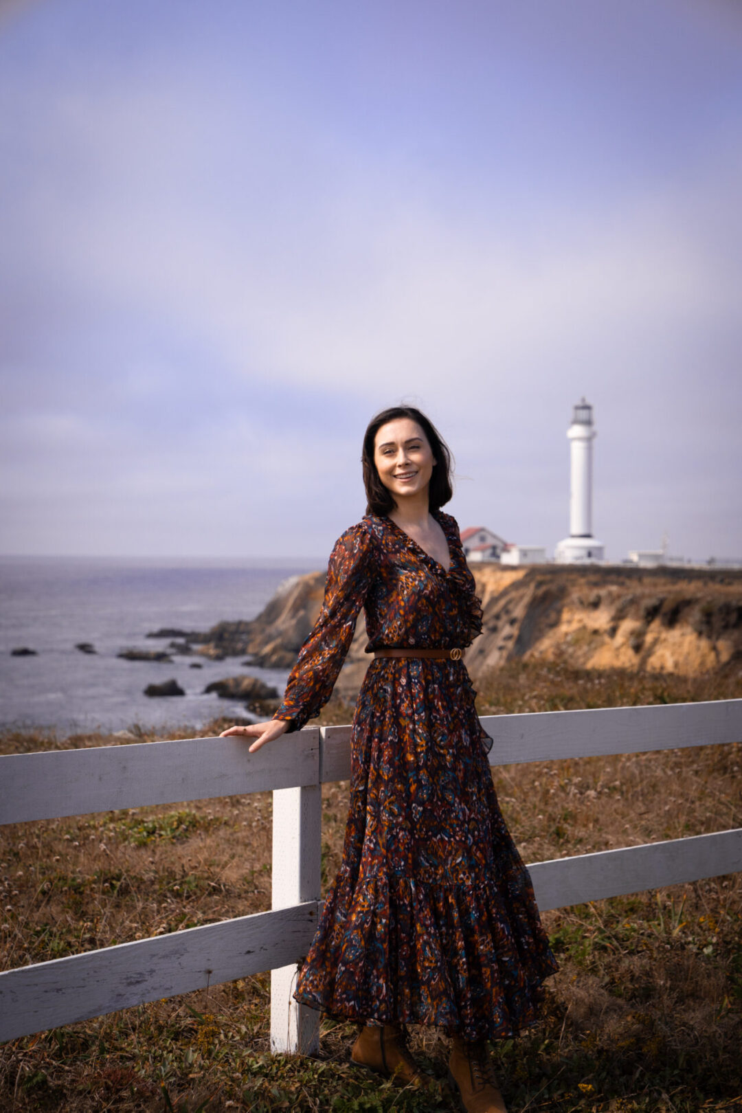 Travel Blogger Jordan Gassner leaning against a white wooden fence adn smiling at a Pacific Ocean lookout point near Point Arena Lighthouse in Northern California