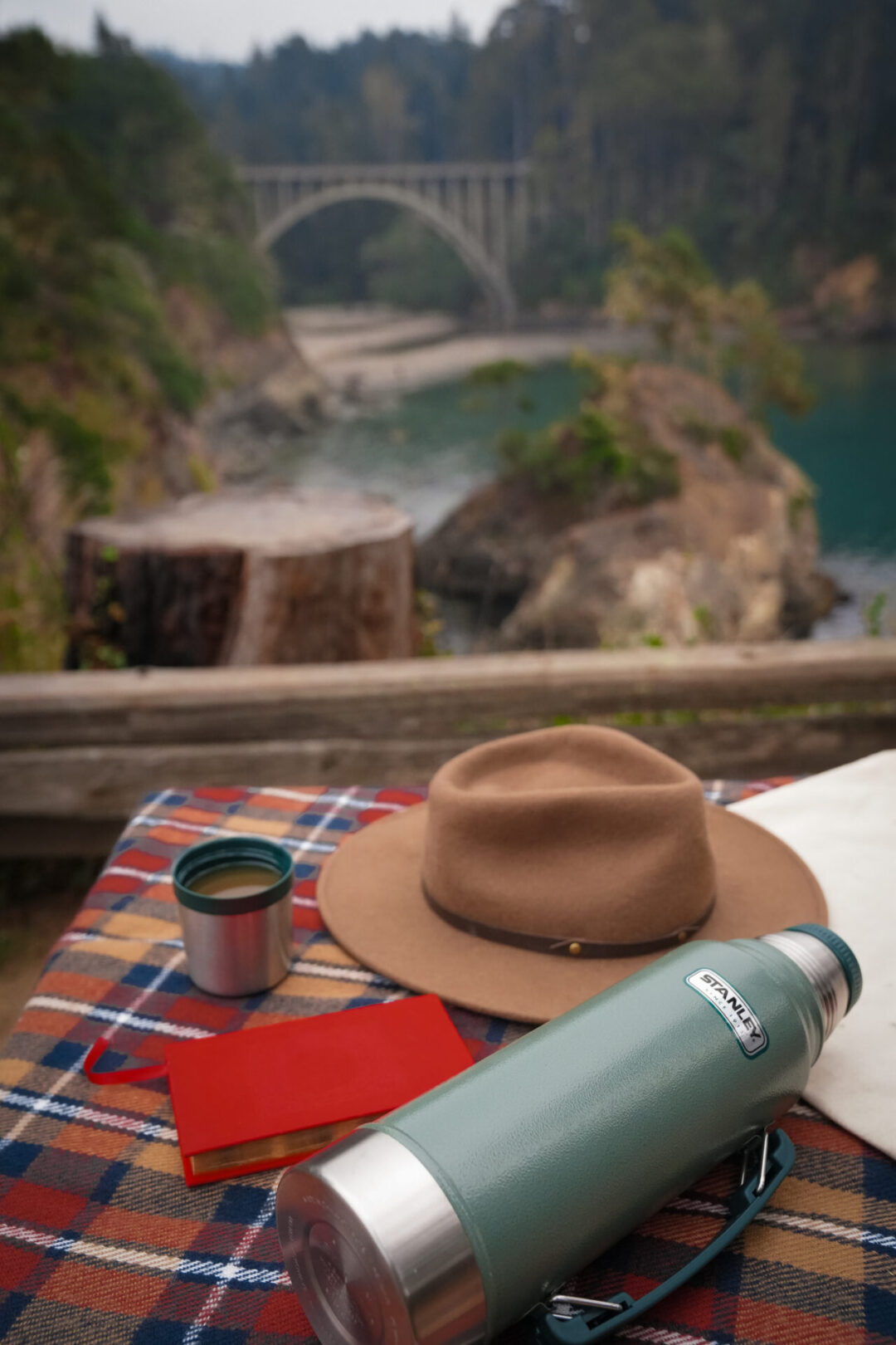 A brown explorer hat, Stanley thermos, red book laying on a red, blue and copper plaid scarf overlooking Russian Gulch State Park in Mendocino, California