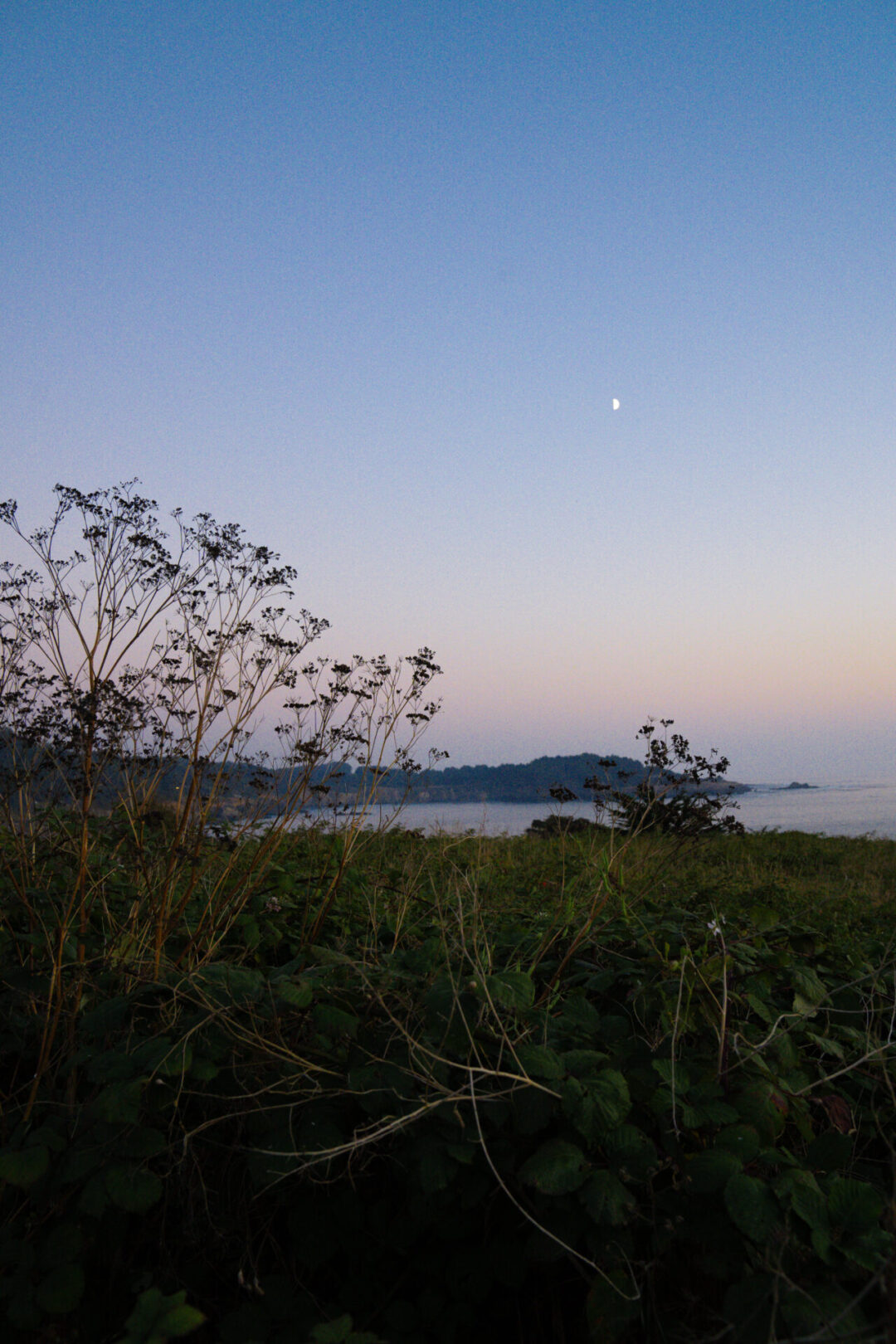 A crescent moon rising over the calm ocean amid purple/orange/blue changing skies in Mendocino, California