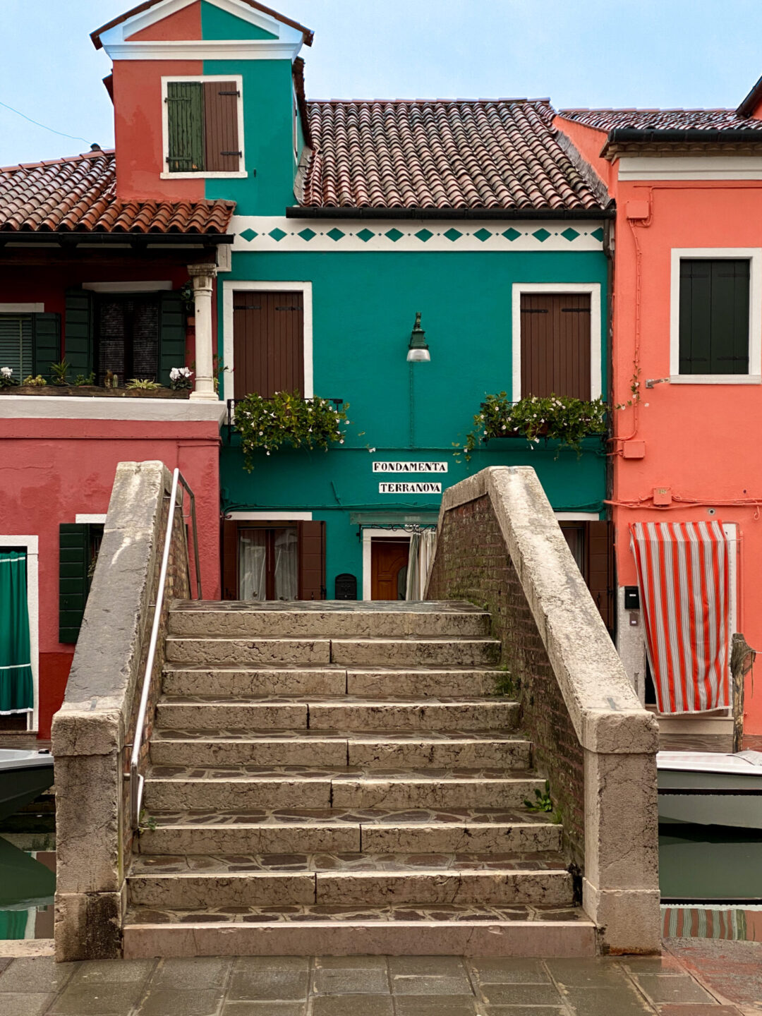 Steps of a bridge leading to three homes: one red, one green and one orange across a canal in Burano, Italy