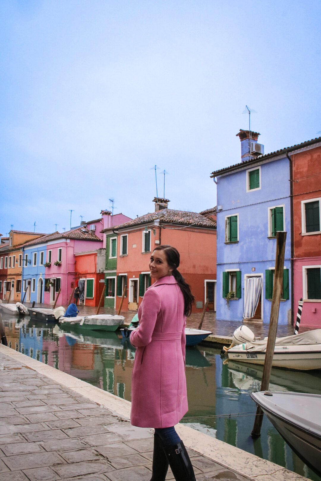 A woman in a pink coat walking along a colorful canal in Burano, Italy near Venice Italy during the winter