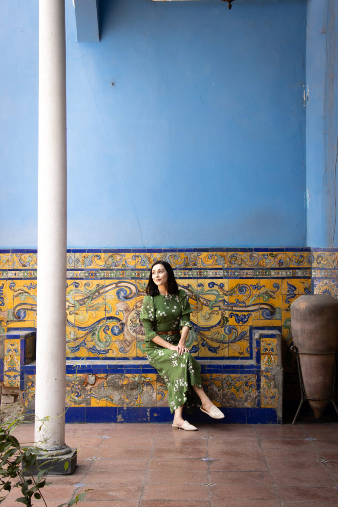 A woman in a green dress sitting on a yellow tiled bench against a blue painted wall in the courtyard of Casa de Larriva in Lima, Peru