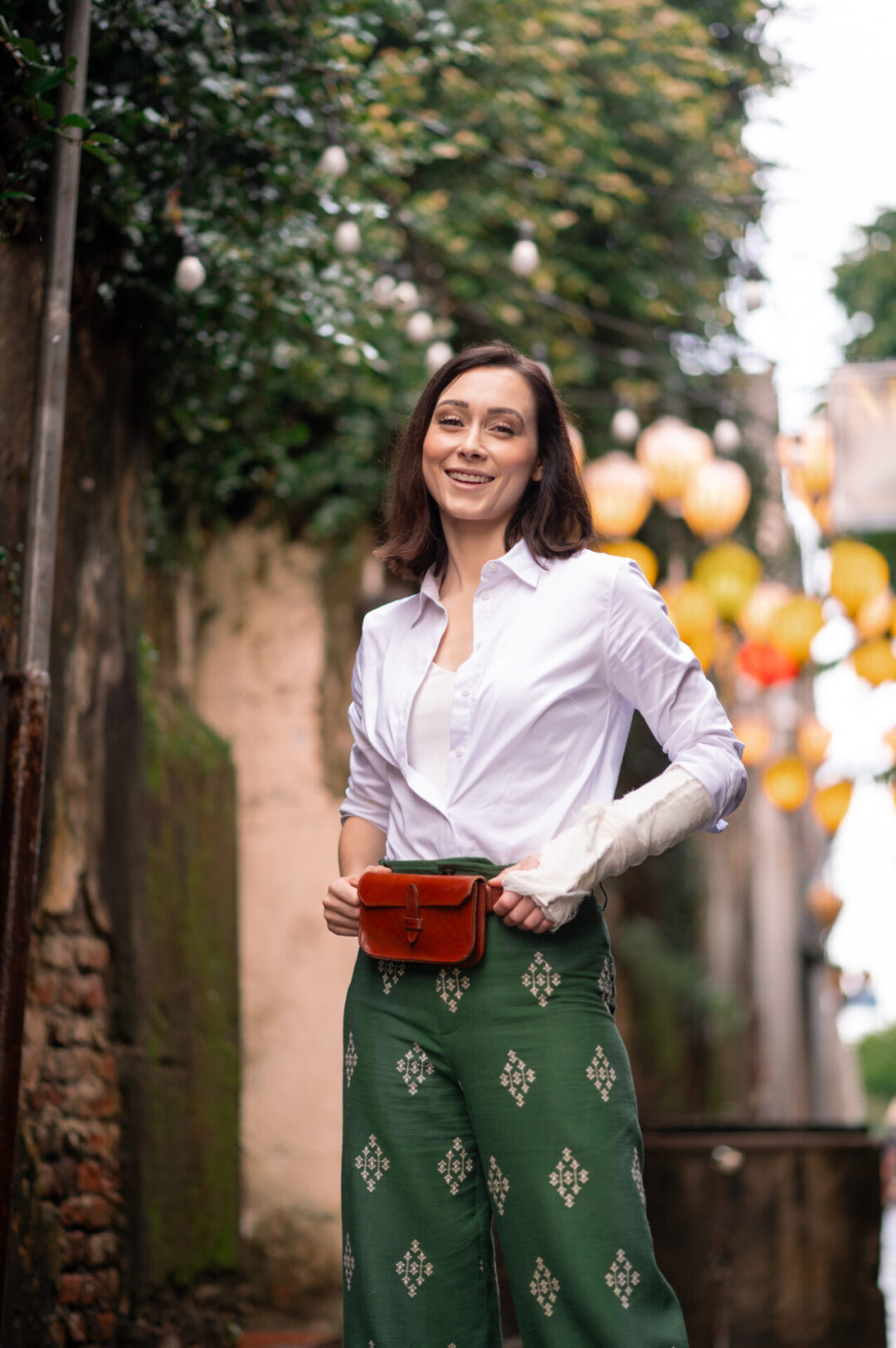 Travel Blogger Jordan Gassner smiling with hands on her brown belt bag and green pants in one of Hoi An's streets, yellow lanterns behind her