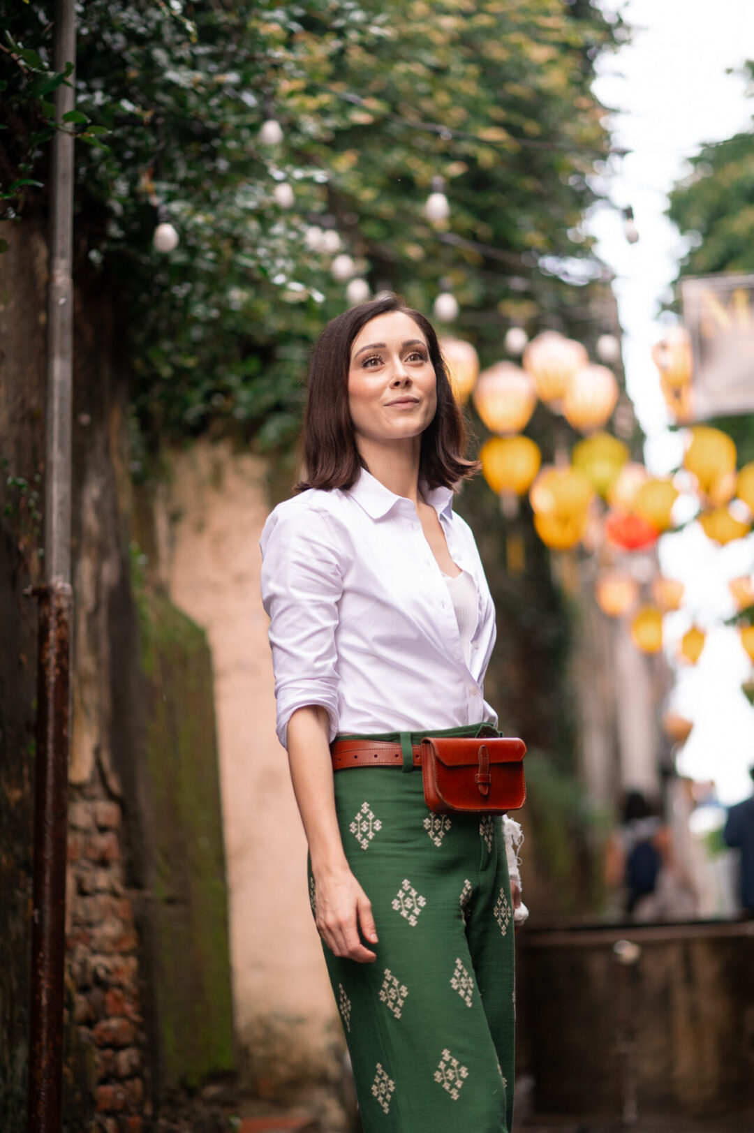 Travel Blogger Jordan Gassner standing in one of Hoi An's streets wearing green pants and a brown belt bag, yellow lanterns behind her