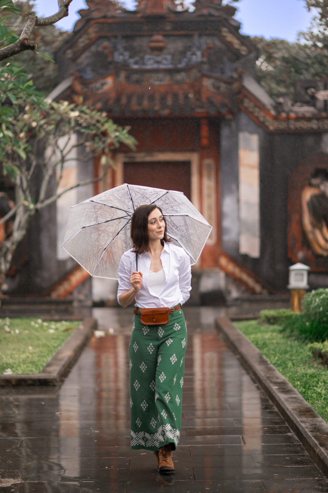 Travel Blogger Jordan Gassner walking underneath an umbrella while wearing green pants and a brown belt bag in front of a temple in Hoi An, Vietnam