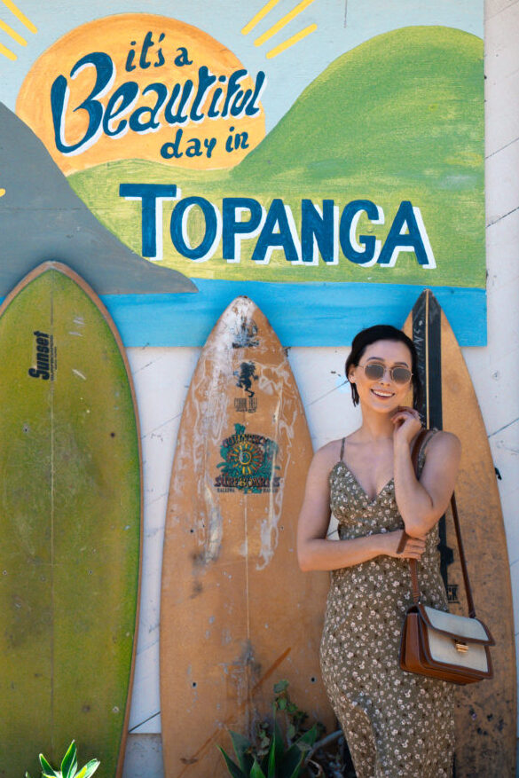 Travel Blogger Jordan Gassner smiling next to a "It's A Beautiful Day in Topanga" mural next to a set of 6 standing surboards inside a Topanga Canyon shopping center