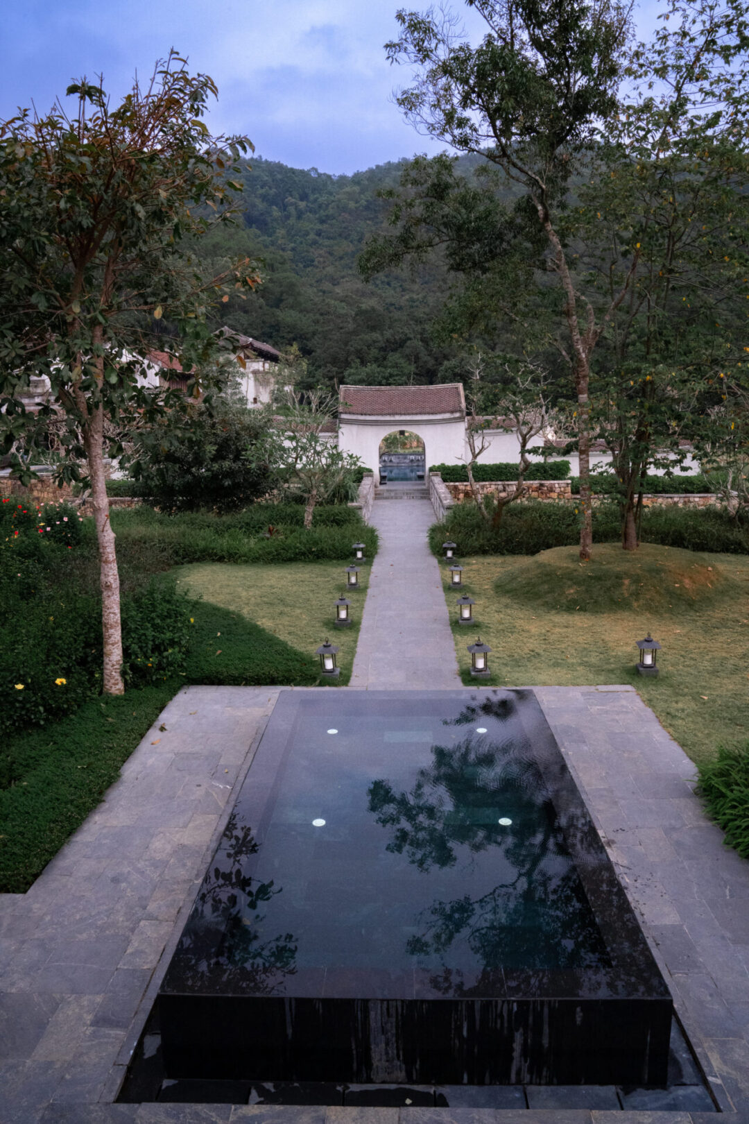 A long empty walkway leading to the pool area at Legacy Yen Tu Resort in Vietnam, surrounded by a natural green garden and towering mountains in the background
