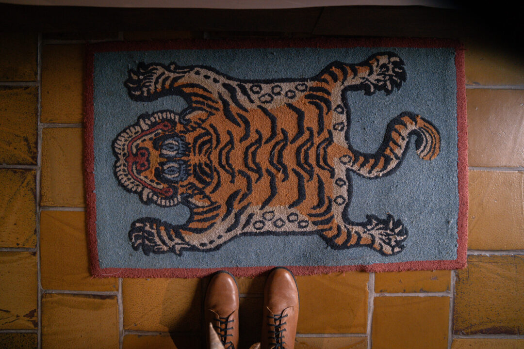 An artistic rendering of a smiling tiger on a vibrant rug laying on a brown tiled floor. A pair of boots just alongside.