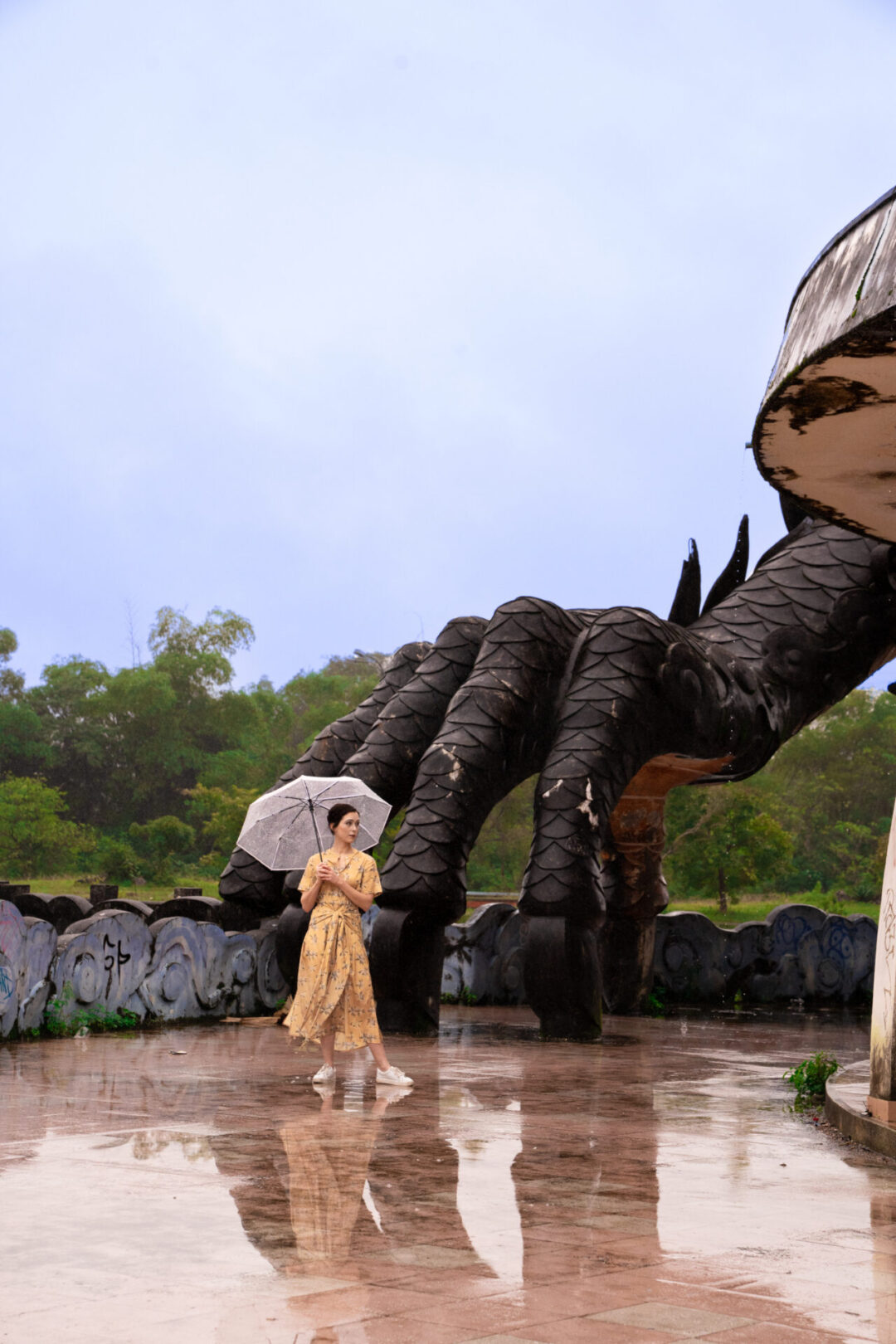 Travel Blogger Jordan Gassner standing with an umbrella near a giant dragon claw that is helping hold up a dragon-like structure at Ho Thuy Tien Abandoned Waterpark in Hue, Vietnam