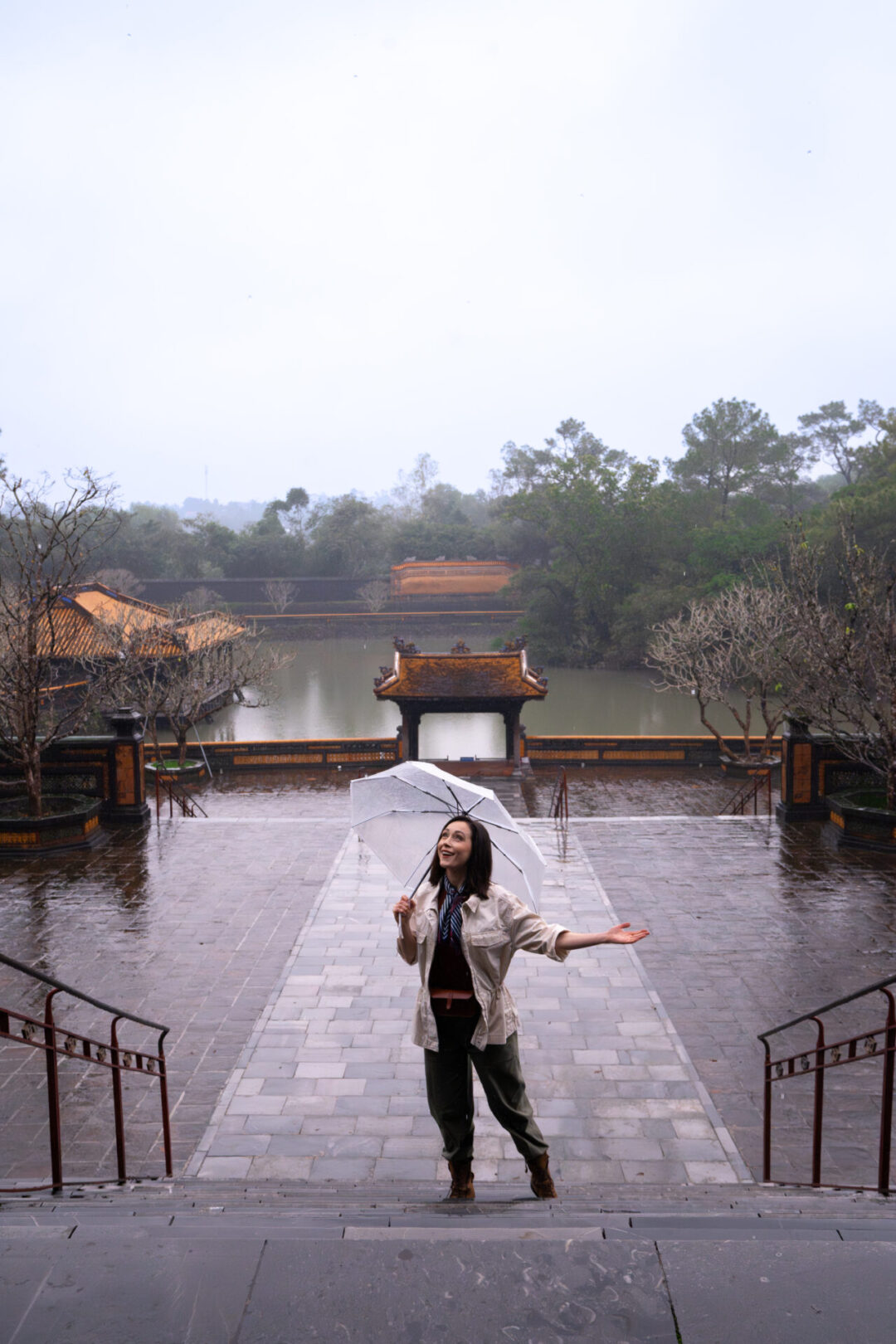 Travel Blogger Jordan Gassner climbing a staircase outside of the Tomb of Tu Duc, holding out her hand from under an umbrella to catch raindrops in Hue, Vietnam