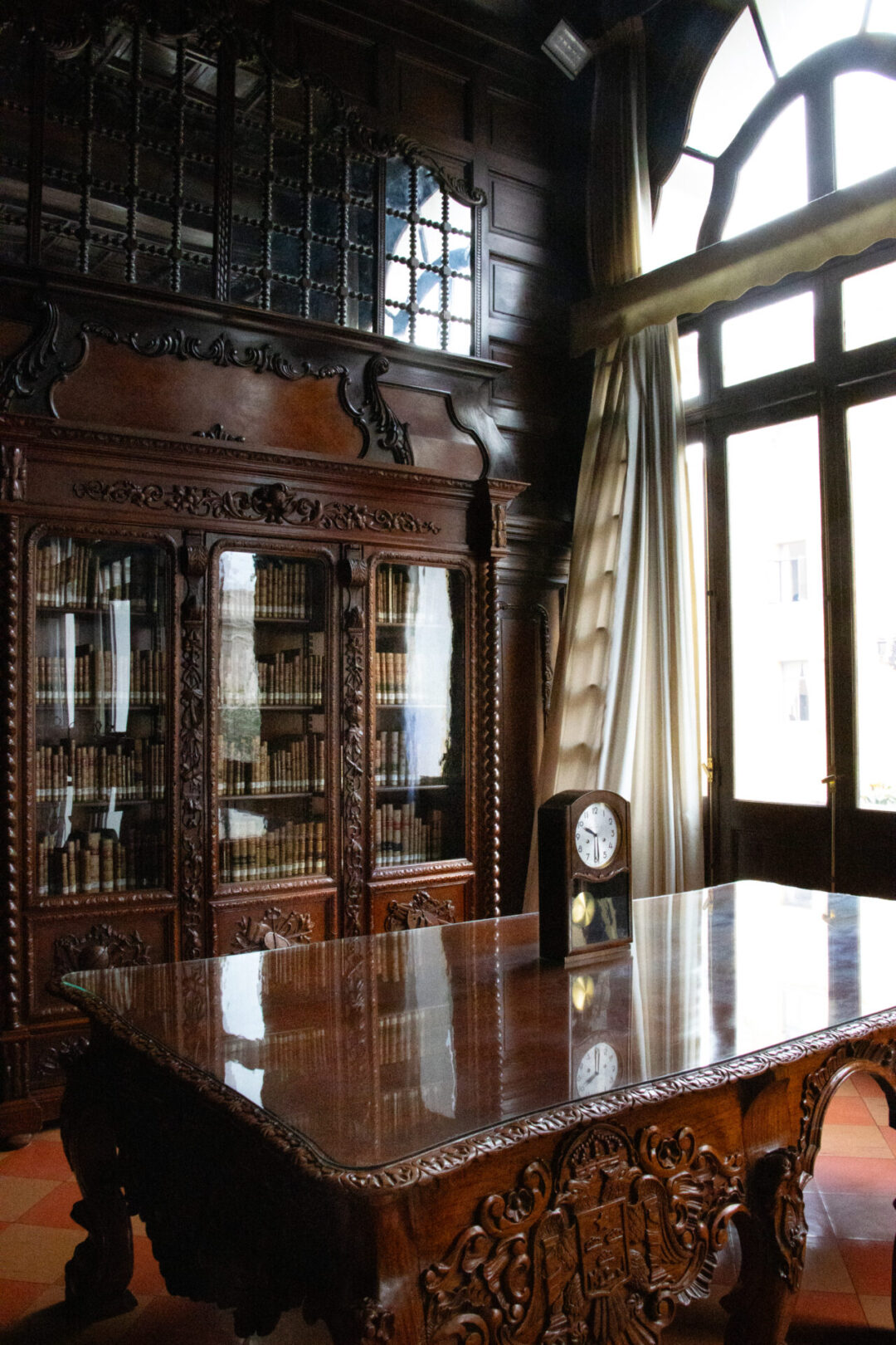 A vintage clock sitting on top of a intricately carved wooden table near an equally exquisite bookshelf inside Lima's Municipal Palace Library