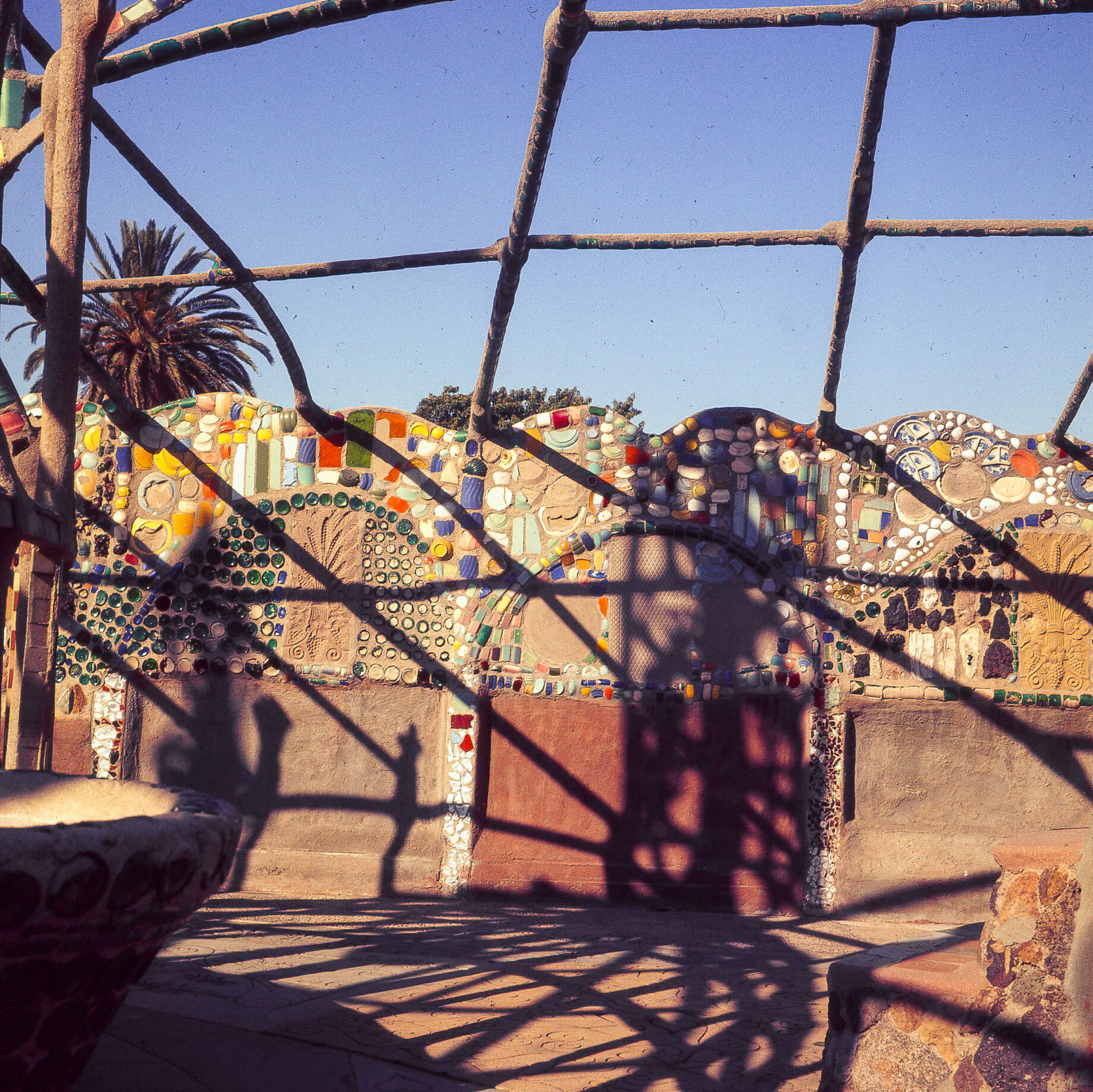 A concrete and metal netted structure above a wall decorated with colorful glass shards in Los Angeles