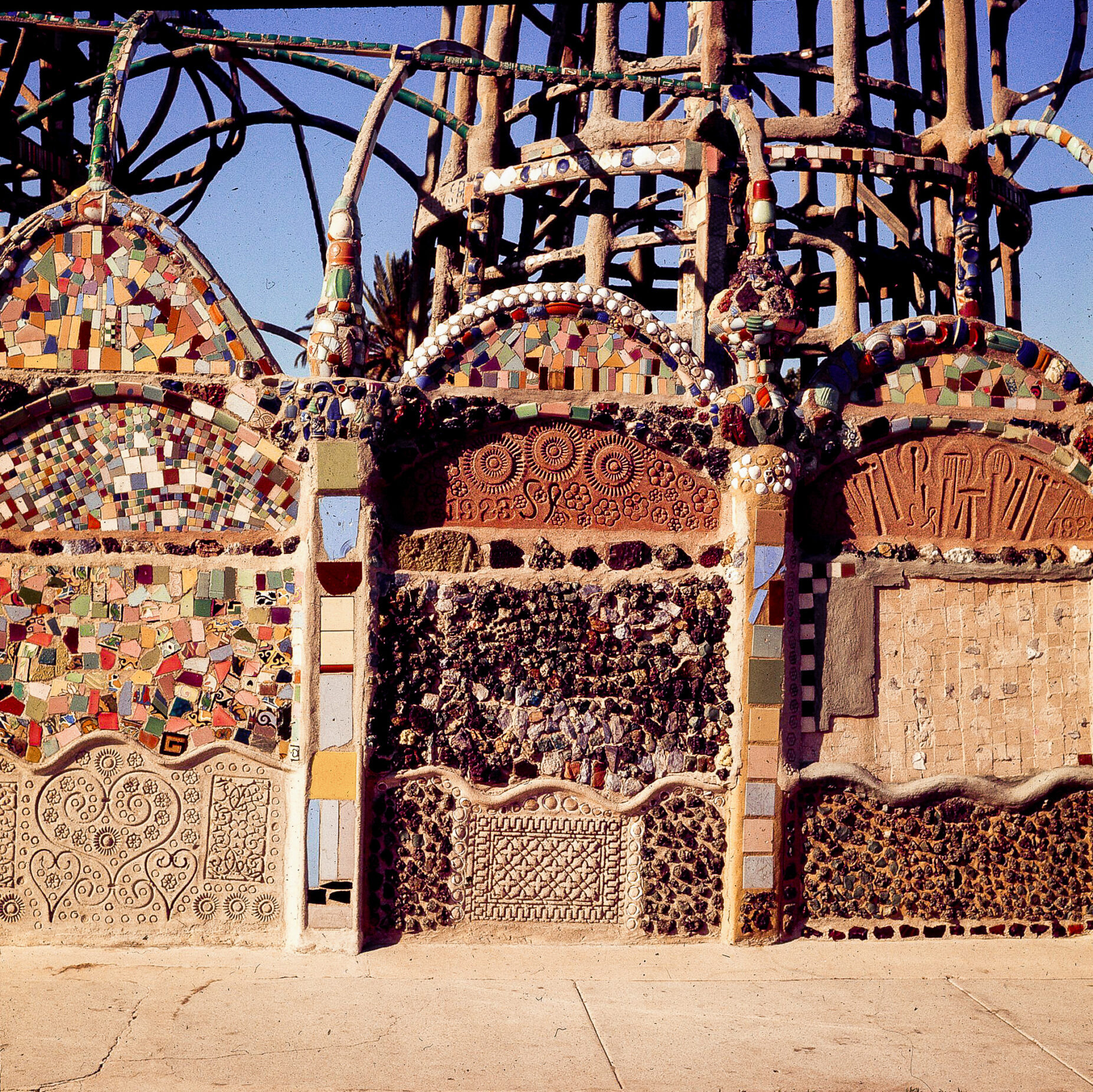 A concrete and metal wall decorated with Tile and glass chards in front of Simon Rodia's Watts Towers in Los Angeles