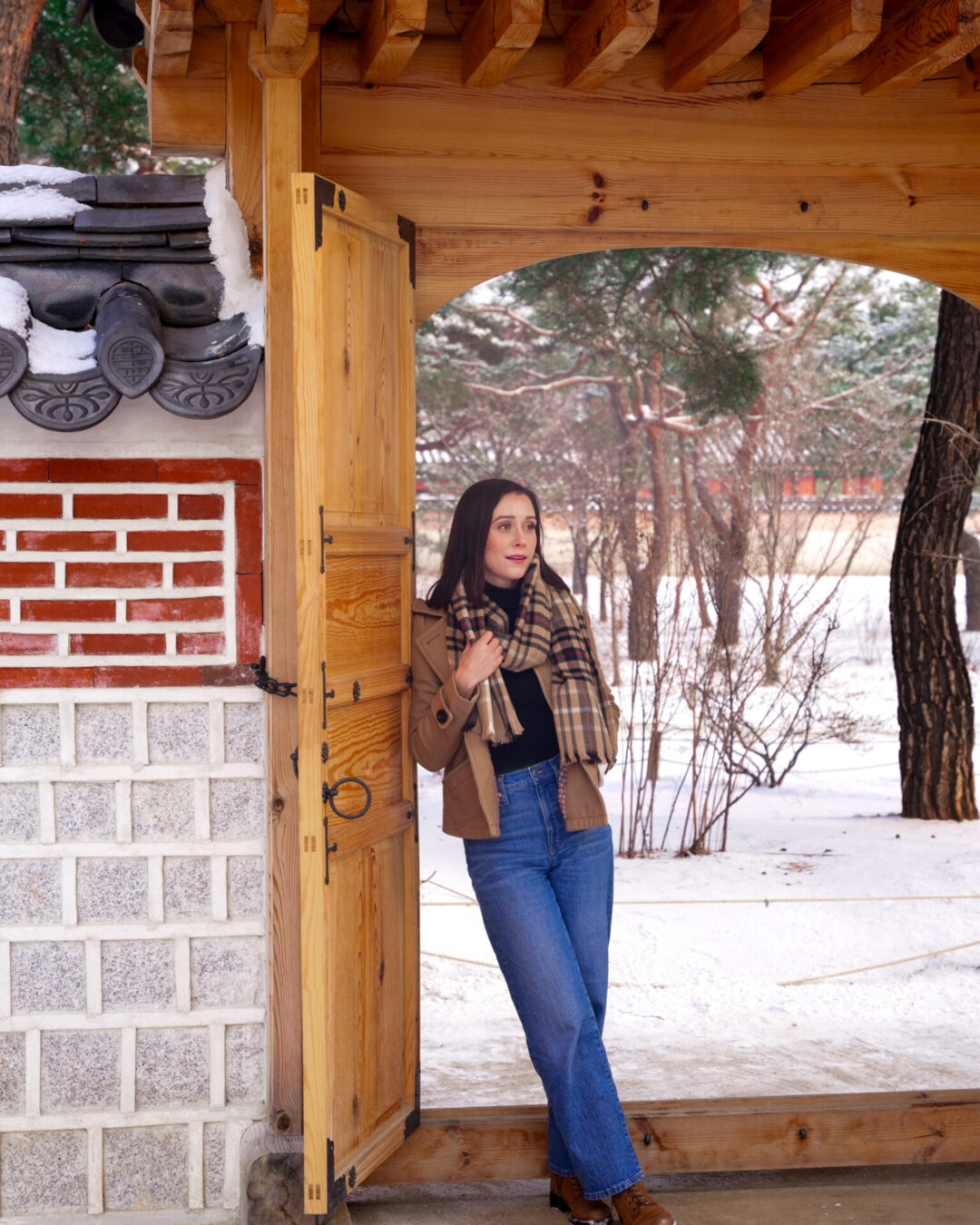 Travel Blogger Jordan Gassner grasping at her scarf while leaning against a gate in between Geonchunmun Gate and Gyejodang Hall on a snowy day