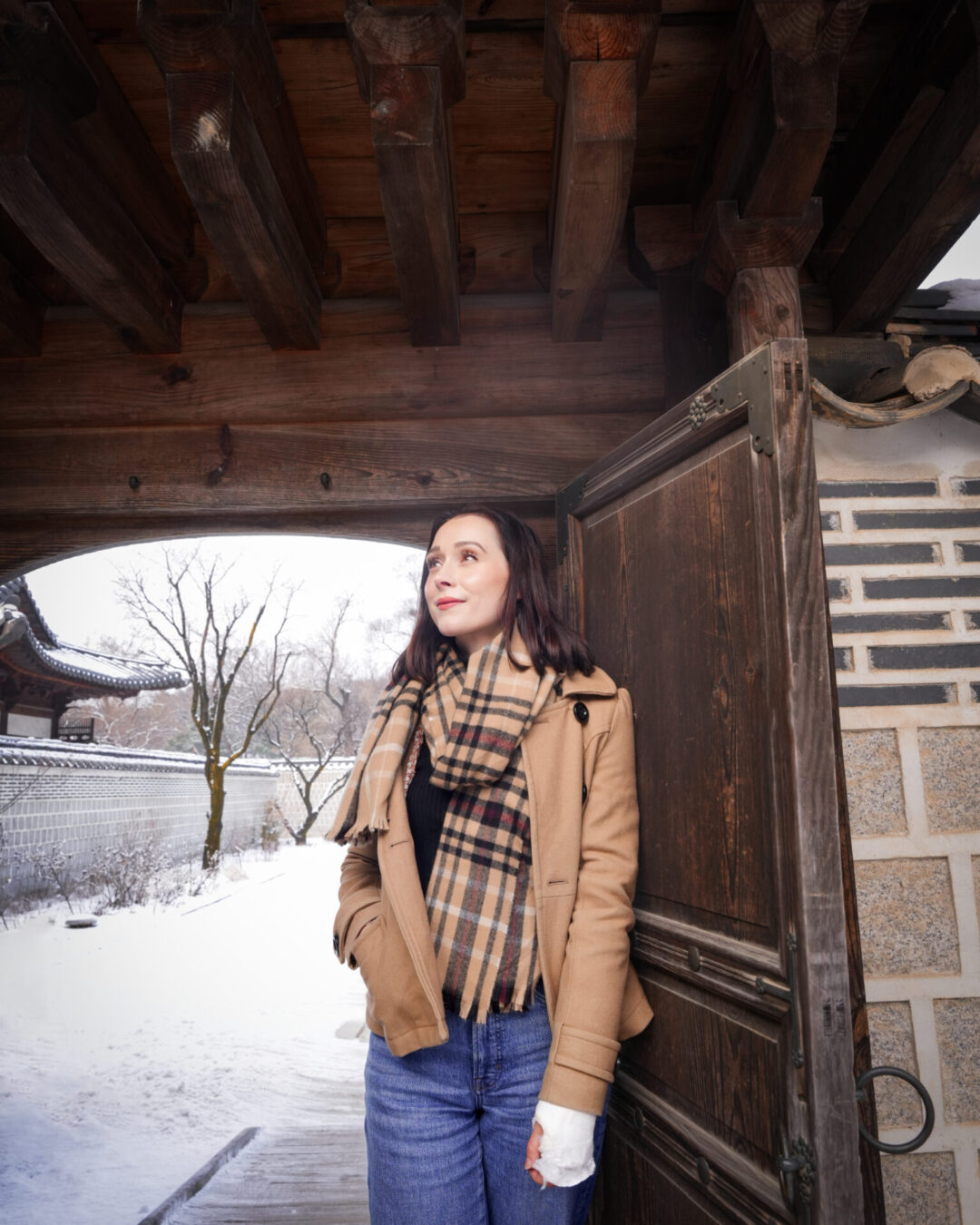 Travel Blogger Jordan GAssner standing at a dark wood gate while bundled in a light brown coat and her arm in a cast on a snowy day in Seoul, South Korea