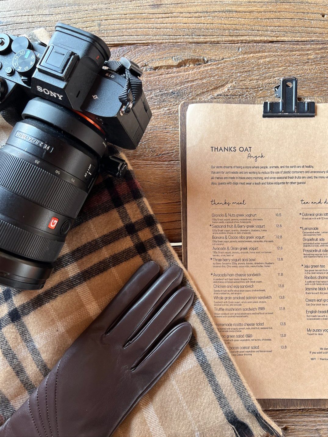 A Sony camera sitting next to a brown leather glove and on top of a scarf near a Thanks Oat Anguk cafe menu in Seoul, South Korea