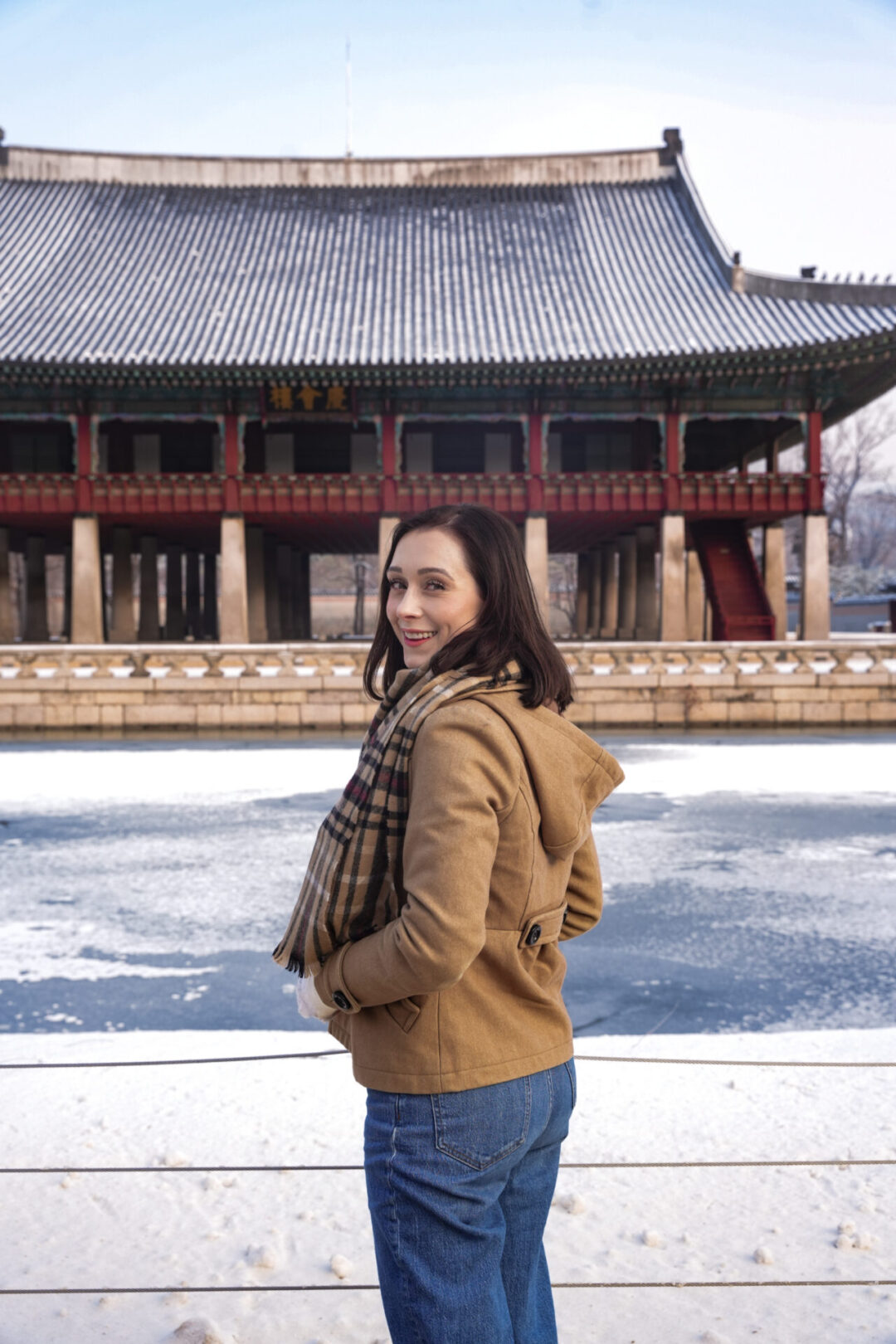 Travel Blogger Jordan Gassner smiling over her shoulder in front of an icy and snow covered pond at Gyeonghoeru Pavilion inside Gyeongbokgung Palace