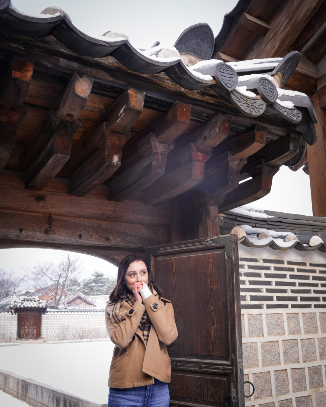 Travel Blogger Jordan Gassner blowing warm air into her hands at a gate leading up to Heungbokjeon Hall on a snowy day inside Gyeongbokgung Palace
