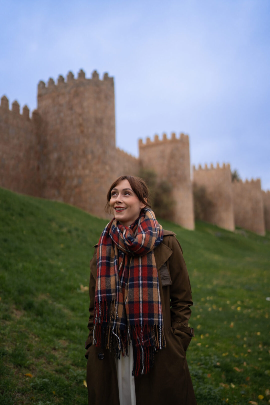 Travel Blogger Jordan Gassner wearing a scarf and looking up at the medieval walls of Avila from a green field in Central Spain