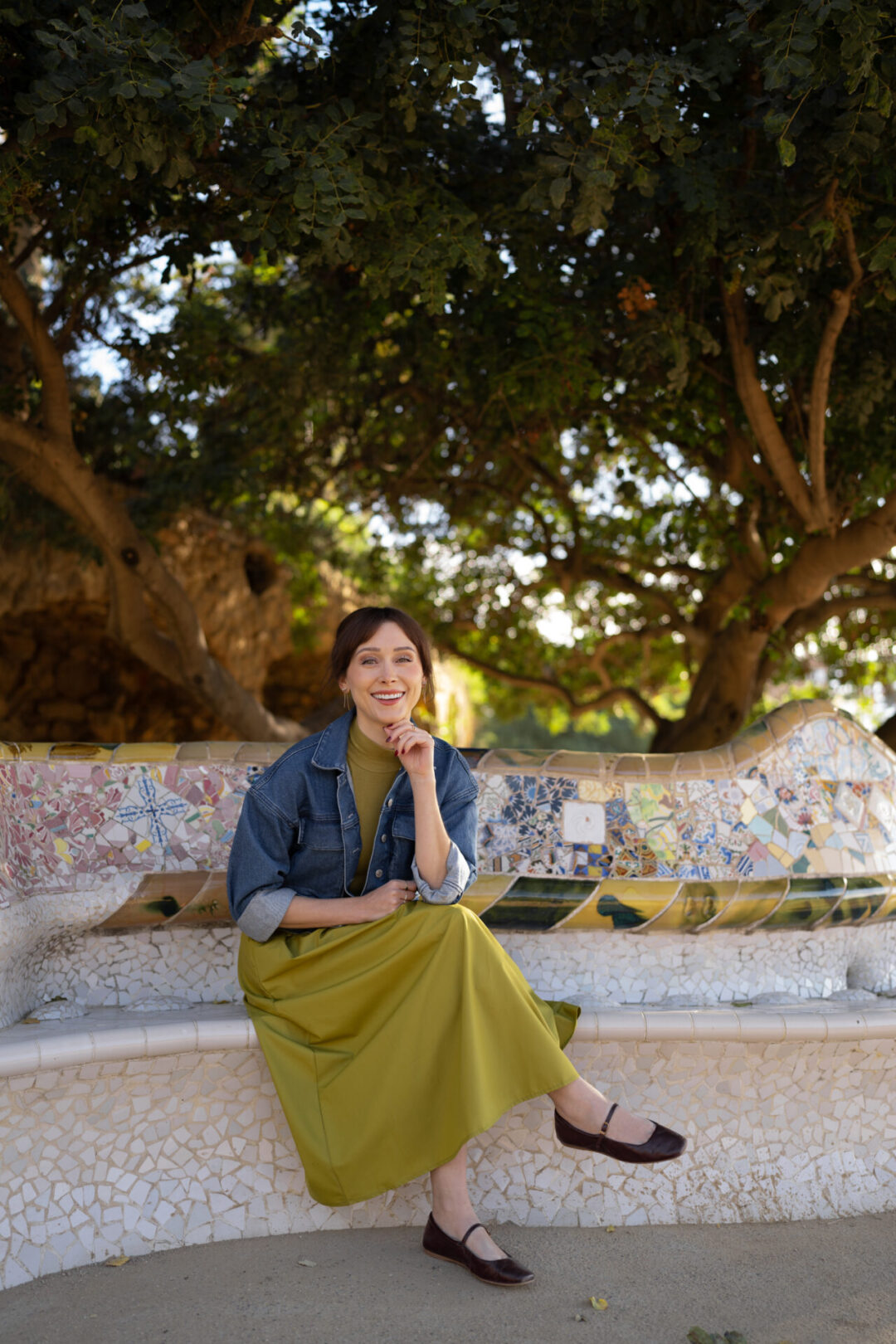 Travel Blogger Jordan Gassner smiling while wearing a green dress and sitting on a mosaic bench in Park Guell in Barcelona, Spain
