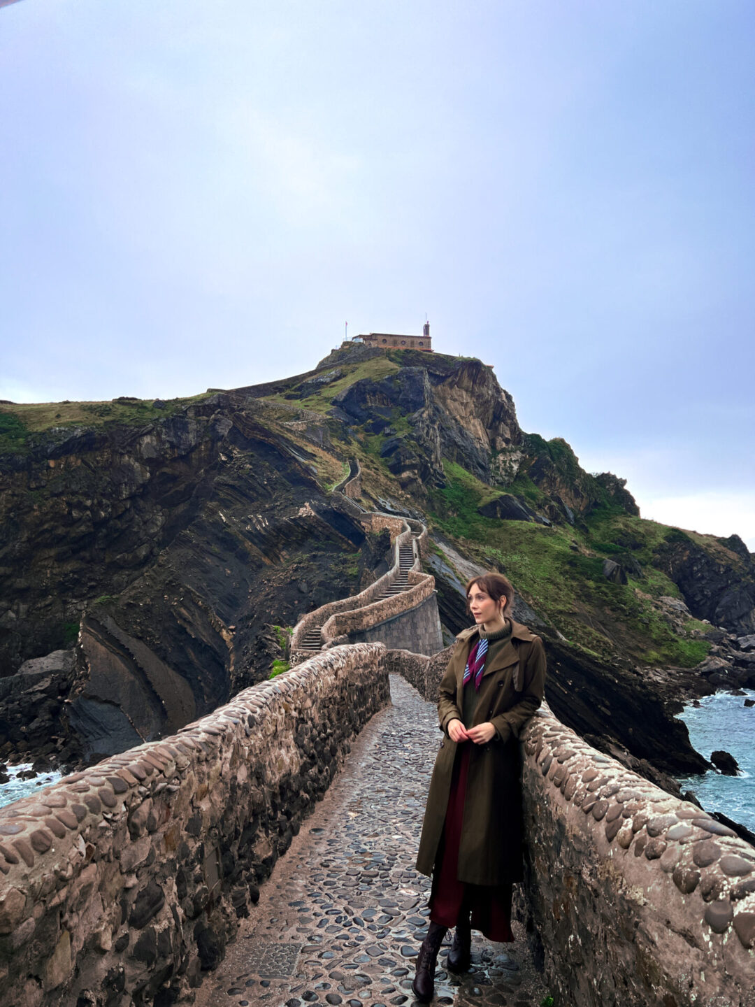 Travel Blogger Jordan Gassner leaning against the stone bridge on a rainy day at San Juan de Gaztelugatxe near Bilbao in Spain