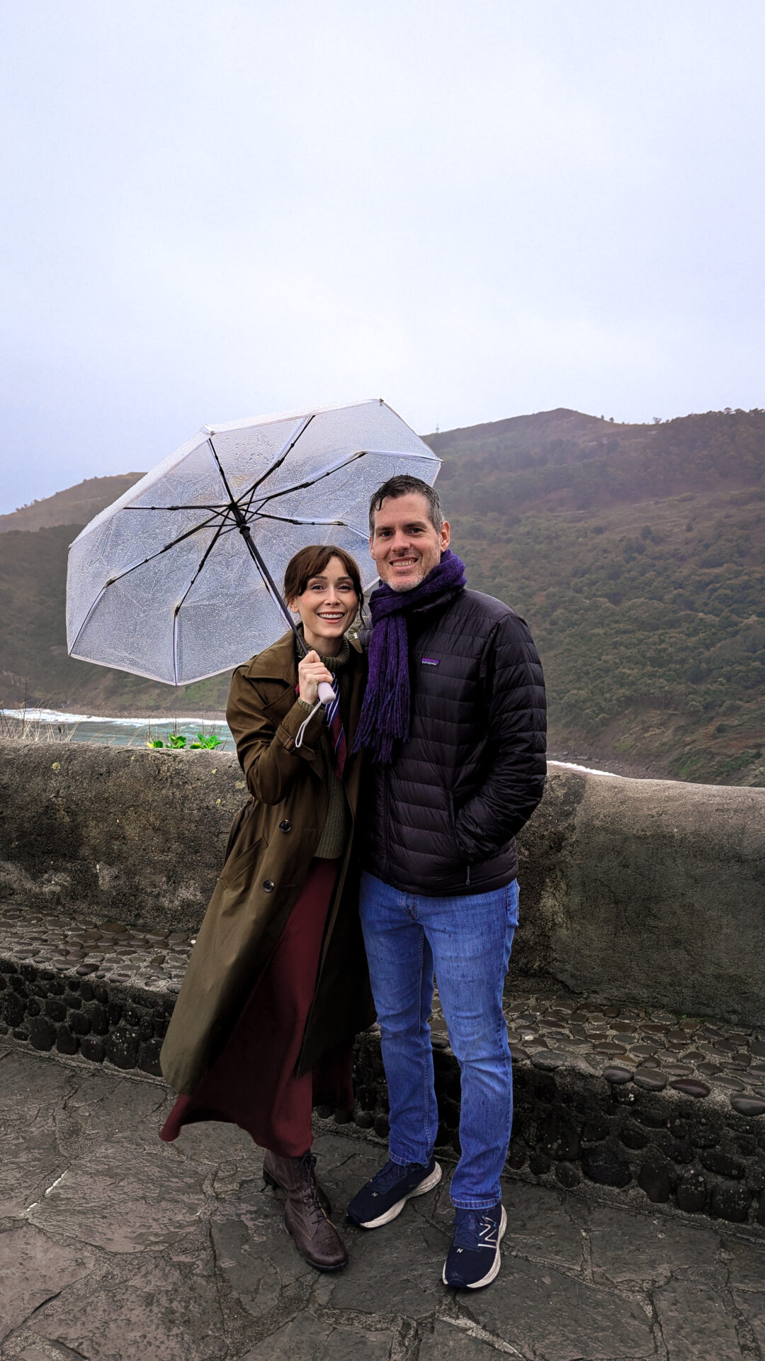 Travel Blogger Jordan Gassner and her partner smiling under an umbrella on a rainy day from the summit of of San Juan de Gaztelugatxe