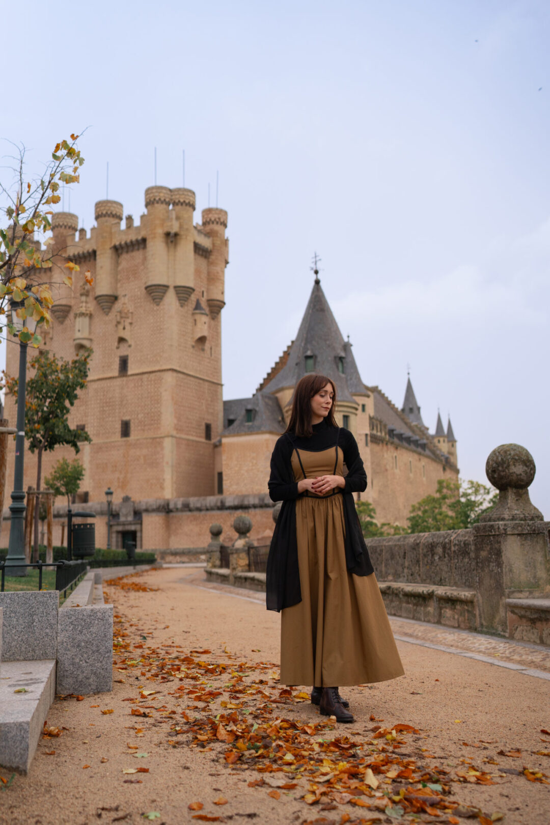 Travel Blogger Jordan Gassner wearing a long dress and looking down at the orange fall leaves near her feet in front of Segovia's Alcazar in Central Spain