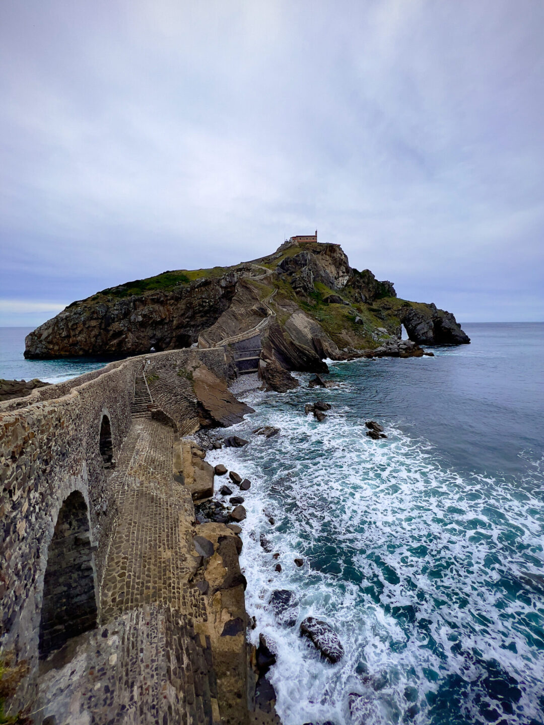 Visiting San Juan de Gaztelugatxe - Waves leading up to the stone bridge at San Juan de Gaztelugatxe on a rainy day in Spain