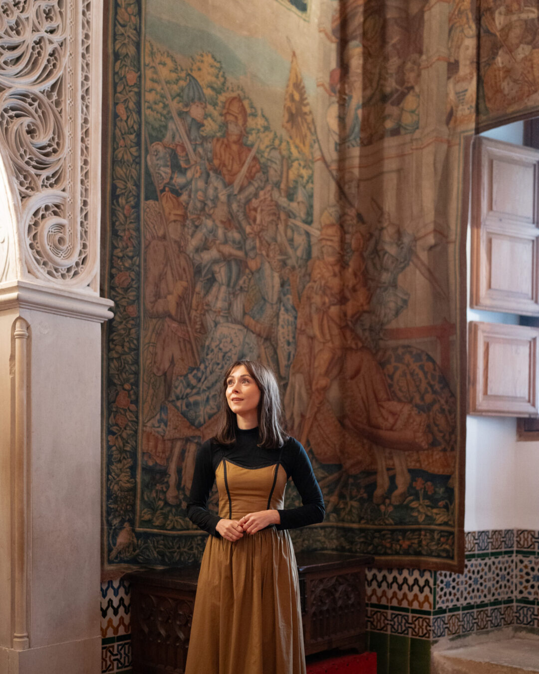Travel Blogger Jordan Gassner wearing a long dress and smiling in front of a wall decorated with a medieval tapestry inside the Alcazar in Segovia, Spain