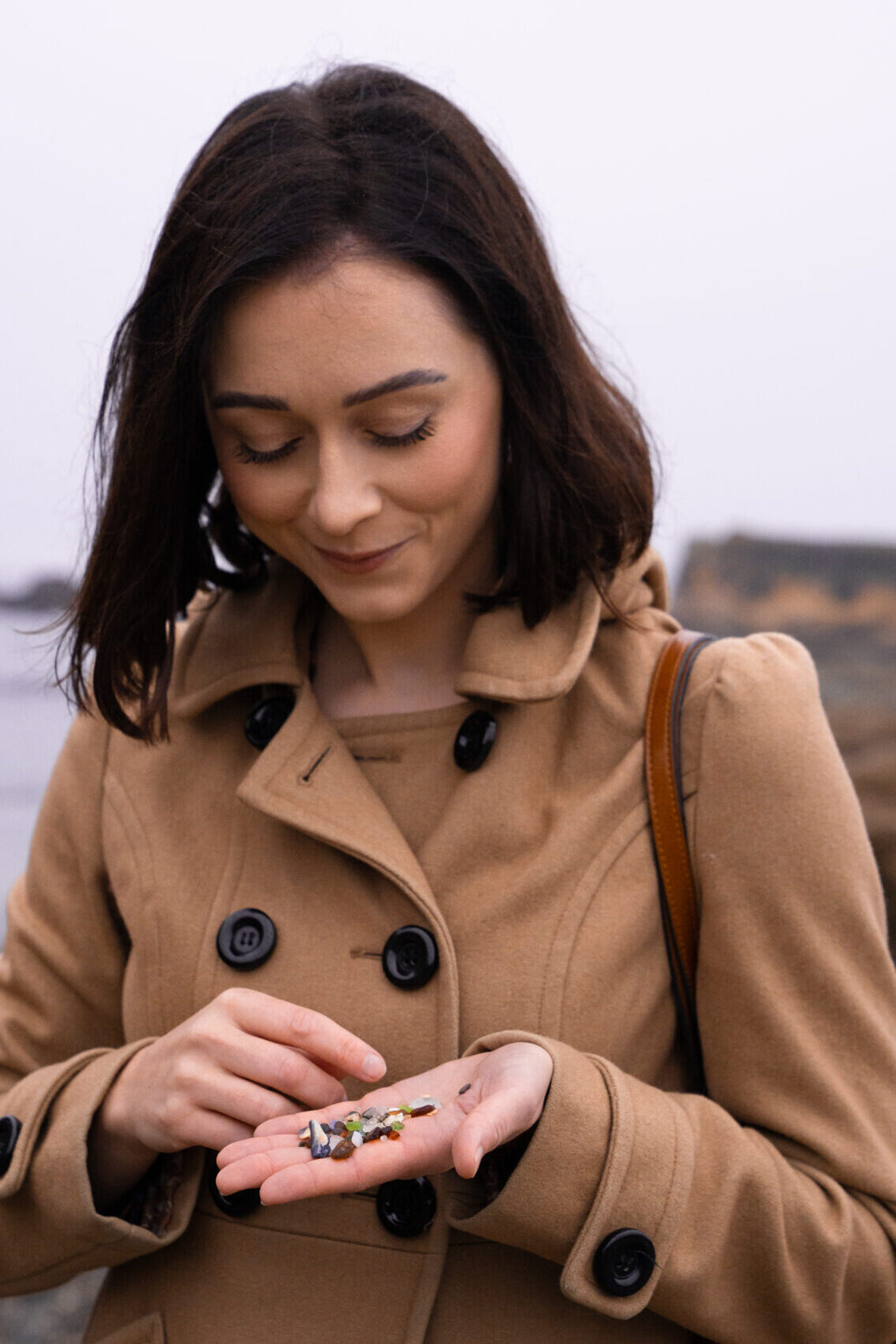 Travel Blogger Jordan Gassner smiling while looking down at some glass-sand in her hand from Glass Beach in Fort Bragg, California