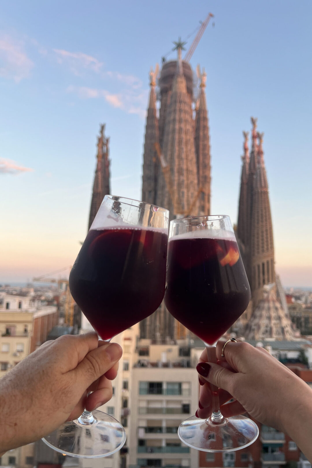 Traveling with a partner after solo traveling - a man's and a woman's hands cheers'ing their sangria glasses on the rooftop overlooking Sagrada Familia in Barcelona, Spain