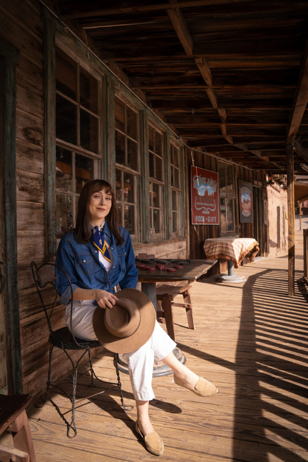 Travel Blogger Jordan Gassner holding her western hat while sitting in front of a vintage wooden table with a chess set at Calico Ghost Town in California