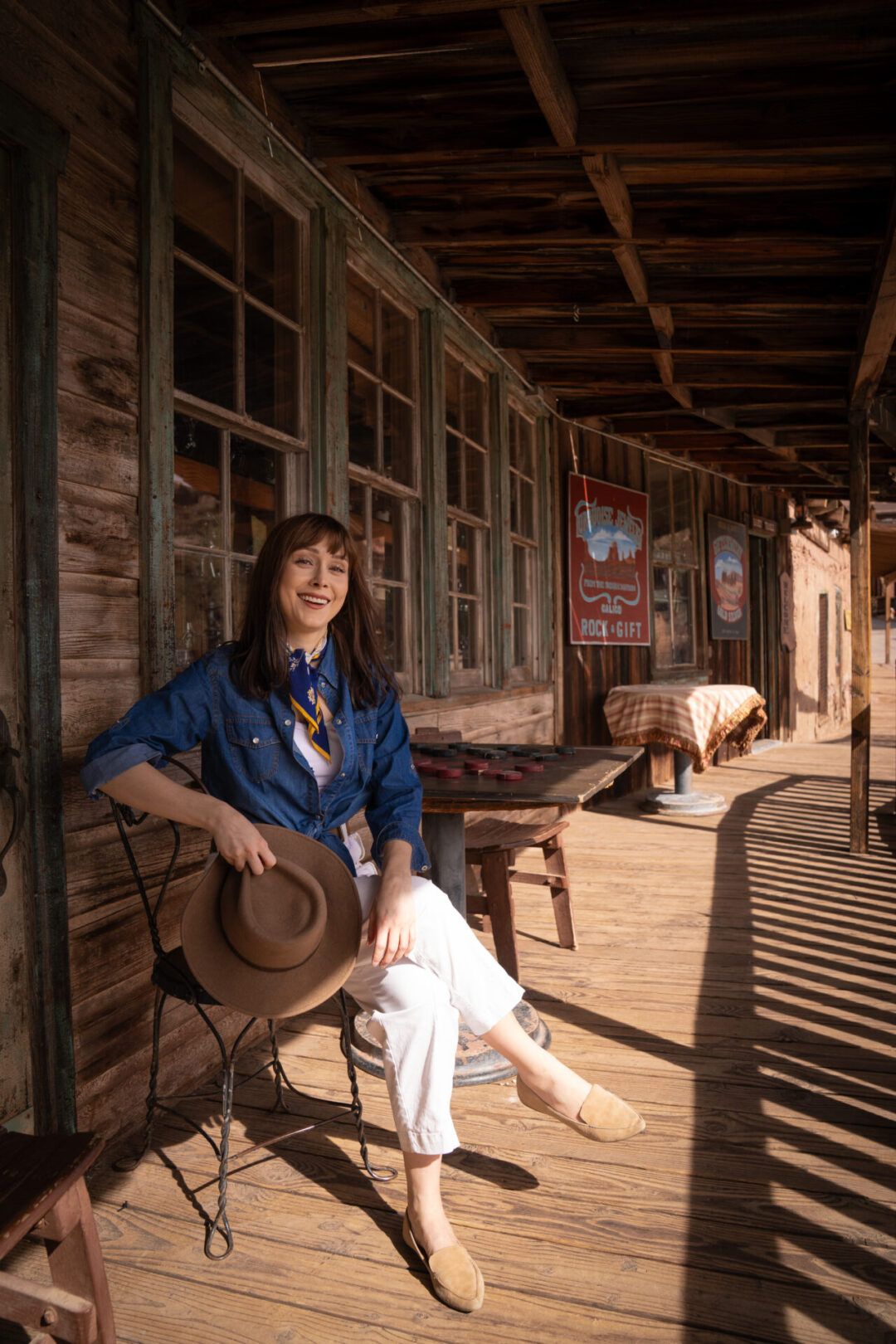 Travel Blogger Jordan Gassner holding her western hat while smiling in front of a vintage wooden table with a chess set at Calico Ghost Town in California